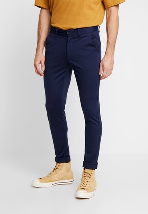 Chinos navy en tissu extensible avec une coupe slim, dotés de passants de ceinture, d'une fermeture à bouton et de poignets retroussés, associés à des baskets montantes beiges.