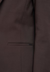 Veste marron foncé cintrée avec une poche filet horizontale et un détail de couture subtil près de la couture latérale.