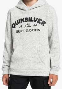Felpa grigia con cappuccio con il testo "Quiksilver 1969 Surf Goods" e logo dell'onda indossata da una persona in pantaloni neri, vista frontale, mani visibili.
