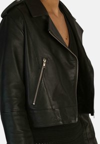 Giacca nera in pelle corta con colletto a punte, cerniere argento e una texture elegante, con tasca laterale con zip e silhouette aderente.