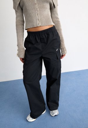 Personne portant un cardigan court côtelé beige et un pantalon cargo noir ample Nike, debout sur un tapis bleu.