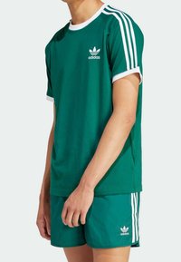 Zelené sportovní tričko s bílými detaily a logem Adidas. Má krátké rukávy, kulatý výstřih a boční rozparky pro volnost pohybu.