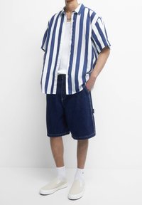 Koszulka krótki rękaw, w paski w kolorze granatowym i białym; zestawiona z ciemnoniebieskimi szortami dżinsowymi; nosi białe skarpetki i beżowe buty wsuwane.