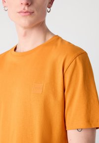 Kortærmet orange t-shirt lavet af bomuld, med en rektangulær logo-patch på brystet og en rund halsudskæring. Glat tekstur.