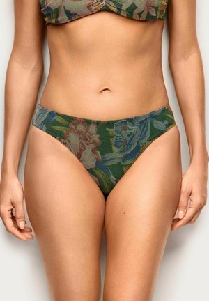 Kobieta w zielonym, kwiatowym dole bikini i dopasowanym topie na jednolitym tle, widoczne tułów i górna część ud.