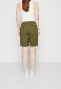 Shorts vert olive en coton avec une coupe droite, dotés de poches arrière et d'un ourlet replié. Portés avec un débardeur blanc et des baskets.
