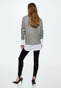Grauer Pullover mit Rippenstruktur, über einem weißen Hemd getragen, kombiniert mit schwarzen Leggings und schwarzen spitzen High Heels. Rückansicht.