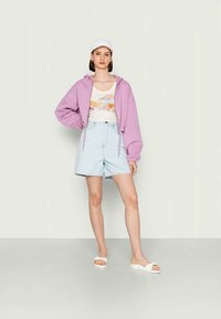 Hoodie oversized lilás, top de alças branco com gráfico, calções de cintura alta azul claro e sandálias brancas. O fundo é neutro.