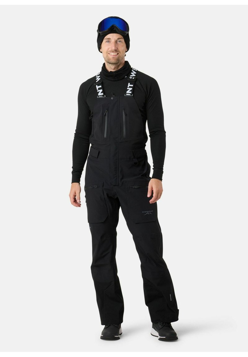 Bib da sci nero isolato con bretelle regolabili, tasche con zip e cuciture rinforzate. Indossato sopra una maglietta nera a maniche lunghe e un berretto.