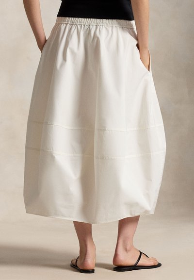 Λευκή μίντι φούστα με ελαστική μέση και πλαϊνές τσέπες, συνδυασμένη με μαύρο τοπ και μαύρα φλατ σανδάλια, σε ουδέτερο φόντο.