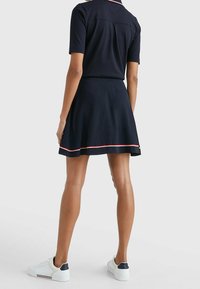 Mujer vestida con una blusa azul marino de manga corta y una falda a juego con ribetes rojos y blancos, combinando con zapatillas blancas, de pie de espaldas.