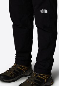 Black waterproof pants with a flexible design and a small white logo on the thigh. Paired with dark green and black hiking shoes with yellow accents.