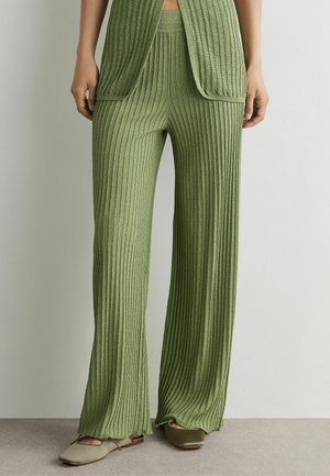 Frau trägt olivgrüne, gerippte Strick-Hose mit weitem Bein, dazu passenden offenen Cardigan und olivfarbene flache Schuhe vor schlichter Wand.