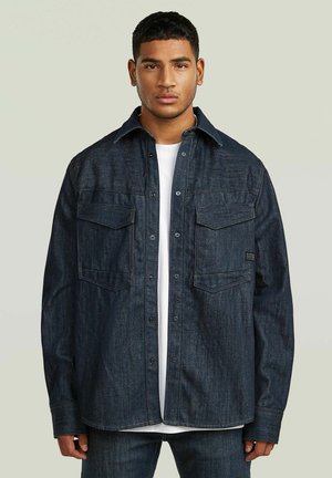 En ung mand står vendt mod fronten iført en mørk denimjakke over en hvid skjorte mod en ensfarvet lys baggrund.