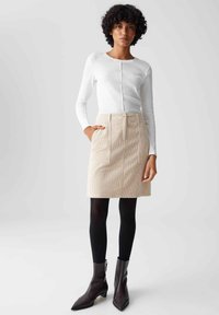 Witte geribbelde longsleeve top, beige gestructureerde minirok met zakken, zwarte panty en puntige bruine enkellaarzen.