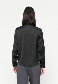 Blusa preta de cetim com mangas longas, textura suave, punhos abotonados e bainha arredondada. Usada sobre calças cinzas, vista de costas.