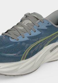 Chaussure de course bleue et grise avec lacets gris, une semelle blanche coussinée étiquetée "NITRO," et une bande latérale jaune et grise.