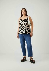 Woman standing wearing black and white patterned sleeveless top, blue jeans, black platform sandals, and a white shoulder bag.