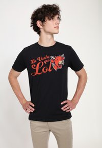 Černé bavlněné tričko s grafikou červené krávy s náušnicemi a textem "La Vache qui Lol" v hravém oranžovém písmu.