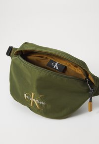 Marsupio in tessuto verde oliva con ricami dorati, apertura con zip e interno marrone. Presenta un'etichetta con logo e componenti in metallo.