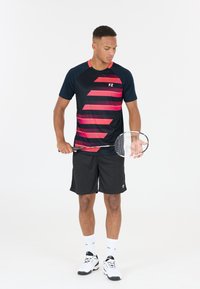 Svarta sportshorts och en marinblå kortärmad skjorta med rosa diagonala ränder. Håller en badmintonracket, stående mot en vit bakgrund.