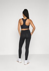 Kvinne med flettehår iført svart sport BH, svarte leggings, hvite sokker og hvite sneakers, stående med hendene på hoftene, vendt bort.