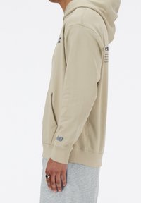 Béžová mikina s kapucí, vybavená přední kapsou, žebrovanými manžetami a šedým logem "New Balance" na rukávu.