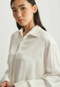 Chemisier blanc à manches longues avec col pointu, fermeture à boutons à l'avant, et une texture légère et lisse. Design ample.