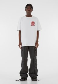 Valge puuvillane T-särk punase graafika logoga rinnal, kombineeritud vabaajastiilis mustade cargo-pükstega, milles on küljetaskud ja nööridega alläär.