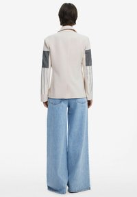 Light beige blazer with striped fabric accents on sleeves, paired with wide-leg blue jeans. Tailored fit with a notched collar.