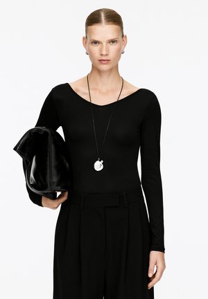 Frau mit nach hinten gekämmtem Haar, die ein schwarzes Langarmoberteil und schwarze Hosen trägt, eine große schwarze Tasche hält und eine runde Anhänger-Kette trägt.