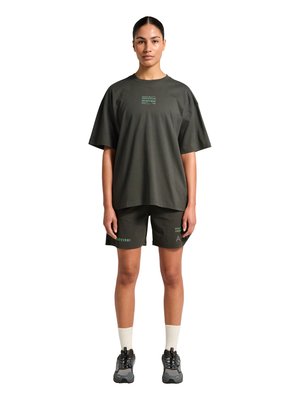 Kvinde står vendt fremad iført mørkegrøn oversized t-shirt og matchende shorts med hvide sokker og grå sneakers på hvid baggrund.