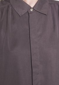 Camicia a maniche corte di colore grigio scuro realizzata in tessuto morbido. Presenta un colletto classico, chiusura con un singolo bottone e pieghe sulle spalle.