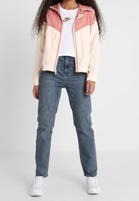 Kvinna iklädd en rosa och krämfärgad zip-up jacka, vit Nike-t-shirt, blå jeans och vita sneakers, som står mot en enfärgad bakgrund.