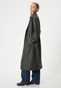 Long manteau gris foncé en matériau semblable au cuir, doté d'un col, de poches latérales et d'une coupe ample. Porté sur un jean à jambes larges.