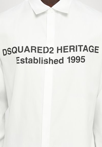 Balta pogām uz augšu t-kreklu ar lieliem melniem burtiem: "DSQUARED2 HERITAGE Dibināts 1995. gadā." Gluda auduma virsma ar klasiska tipa apkakli.