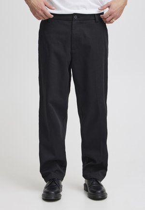 Homme portant un pantalon noir à jambes droites, des chaussures noires à lacets et un t-shirt blanc, tenant la ceinture des deux mains.