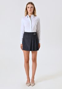 Camicia bianca con bottoni abbinata a una gonna plissettata in denim scuro con dettaglio a fibbia laterale, completata da tacchi beige a punta.