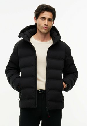 Mand iført en sort dunjakke med hætte over en beige sweater, stående med hænderne i lommerne mod en ensfarvet baggrund.
