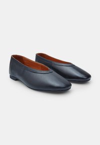 Zapatos planos de cuero negro con punta redondeada, tacón bajo y textura suave. El interior presenta un forro marrón en contraste.