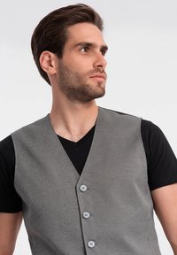 Grau-schwarz karierter Weste aus strukturiertem Stoff mit V-Ausschnitt, fünf Knöpfen, die über einem schwarzen T-Shirt getragen wird.