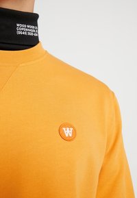 Oranje katoenen sweatshirt met een rond, geborduurd logo in het wit op de borst. Het heeft een geribbelde zwarte col.