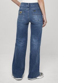 Mėlynos džinsinės kelnės su aukštu liemeniu ir platėjančiomis kojų apačiomis, dvi užpakalinės kišenės, logotipo žymė ir išblukusi tekstūra. Siūtas apačios kraštas su šiek tiek nusidėvėjimo ženklų.