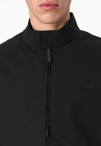 Jachetă neagră cu fermoar, cu guler înalt, dintr-un material neted, având un mic logo brodat negru pe partea stângă și o buzunare cu fermoar.