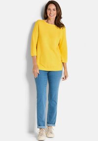 Gelber Strickpullover mit Dreiviertelärmeln, kombiniert mit hellblauen geraden Jeans und beigen Sneakers. Schlichte Gestaltung, weicher Stoff.
