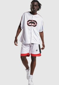 Witte sportieve shirt met zwarte en rode accenten, voorzien van een groot grafisch logo. In combinatie met witte shorts met rode afwerking. Gestructureerde stof, sportief ontwerp.