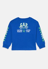 Blaues Langarmhemd aus Baumwolle. Die Vorderseite zeigt "GAP" und Rennflaggen; die Ärmel haben grüne grafische Quadrate; fetter Text enthält die Namen der Charaktere.