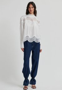 Blouse blanche avec des détails en dentelle le long de l'encolure et de l'ourlet ; coupe ample ; manches longues ; associée à un jean taille haute bleu foncé et des sandales noires.