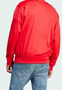 Punainen vetoketjullinen verkkatakki, jossa on sileä pinta, ribbireunat helmassa ja hihansuissa sekä rento leikkaus, yhdistetty vaaleansinisiin farkkuihin.