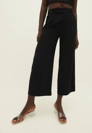 Pantalones negros de pierna ancha y cortos, usados por una persona con sandalias transparentes de tacón bloque, sobre un fondo claro.